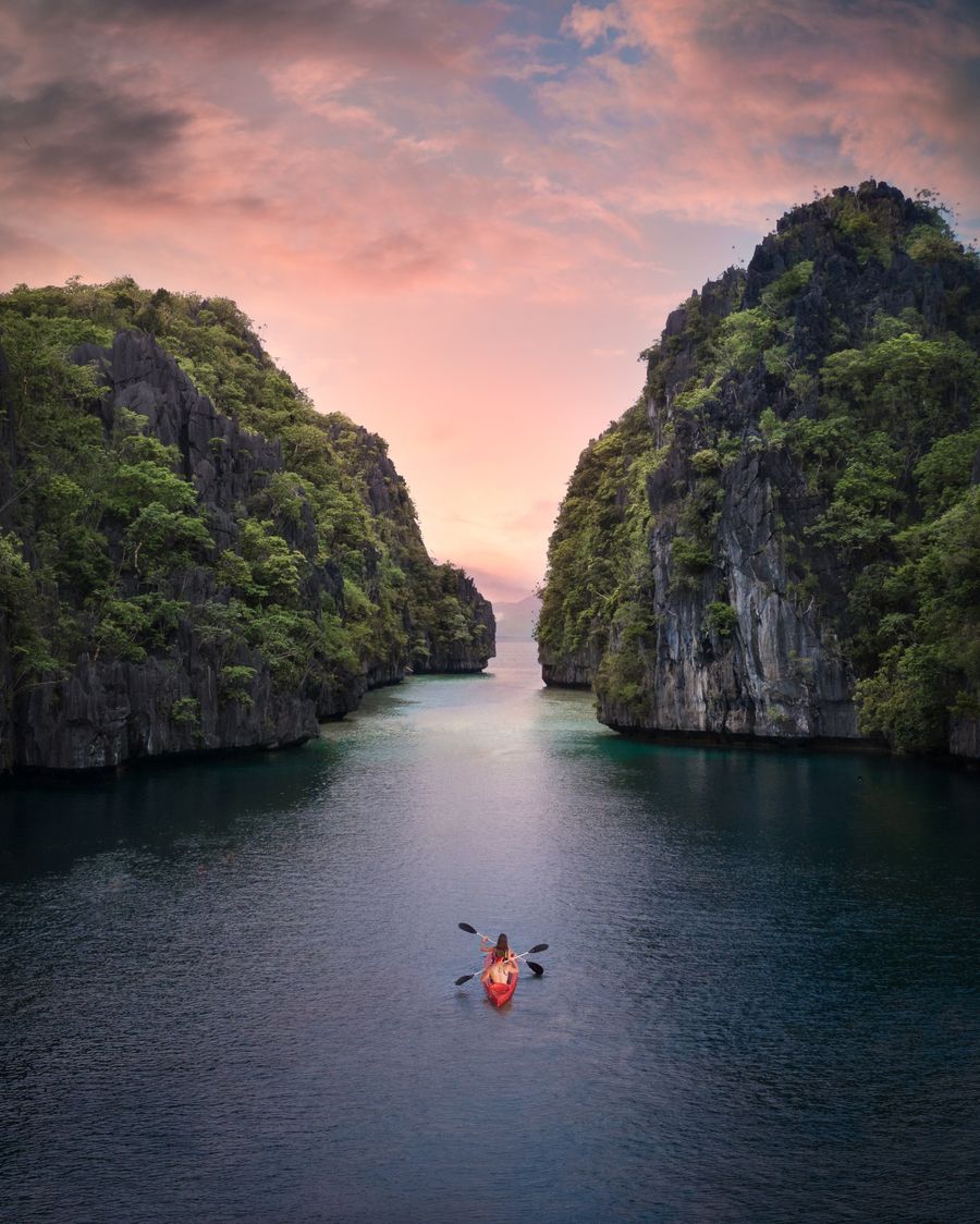 Two people paddling a kayak between towering green cliffs toward open water at sunset